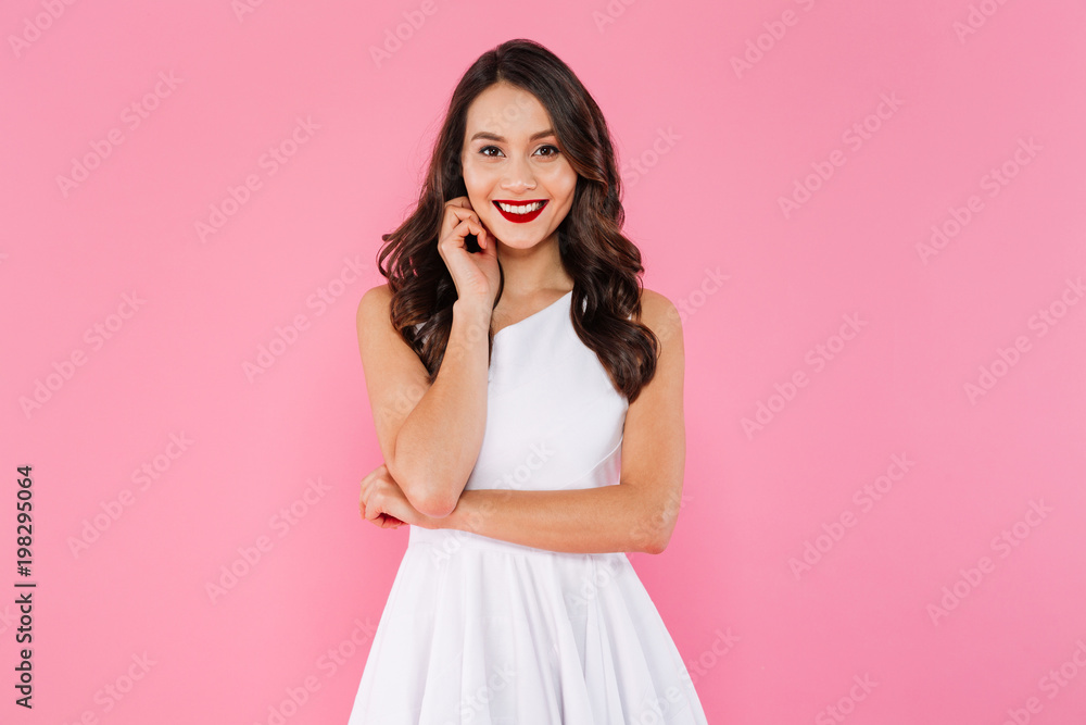 Wall mural Portrait of asian lovely woman with dark curly hair in white dress posing with kind smile, isolated over pink background