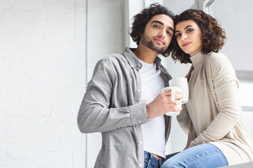 young couple holding cups of coffee and looking at camera at home