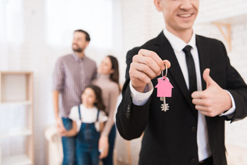 Realtor in suit holds keys with key fob in form of house. Young family looks apartment.