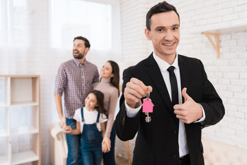 Realtor in suit holds keys with key fob in form of house. Young family looks apartment.