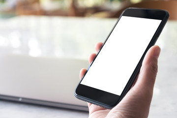 Mockup image of hand holding black mobile phone with blank white desktop screen and laptop on table