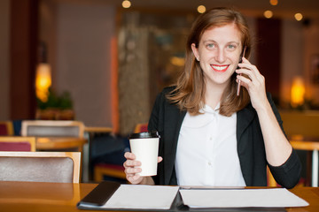 Portrait of cheerful young Caucasian female freelancer drinking coffee, talking on mobile phone,...