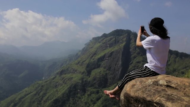the girl is sitting on the cliff and taking photo.