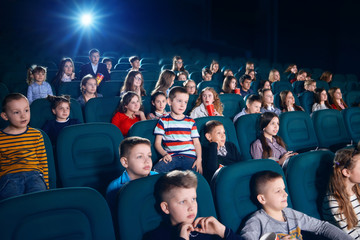 Sideview of children watching movie in the cinema hall. Kids are very emotional, exited and satisfied. Some children eating popcorn or drinking fizzy drinks.