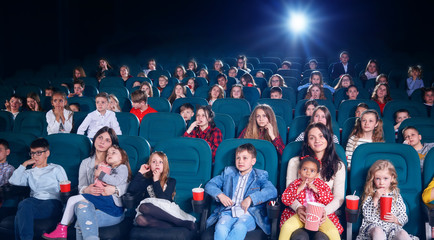 Fototapeta premium photo of cinema hall full of people. Adults and children watching intresting movie,eating popcorn,drinking fizzy drinks. Kids are very emotional : smiling, laughing, looking exited and satisfied.