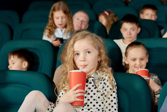 Pretty Girl With Blonde Curly Hair Drinking Fizzy Drink In Cinema. Kid Wearing White Dotted Dress With White Tights. There Are Many Little Children Watching Movie On Background.