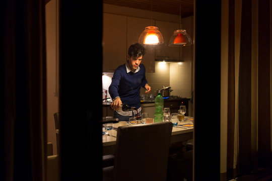 Young man pouring wine in the kichen at night, seen from outside