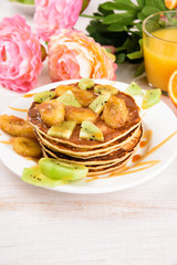 Homemade breakfast on a wooden white table. Stack of pancakes with a grilled bananas and a caramel. Glass of juice, orange, kiwi slices. Pink flowers, flat lay, top view