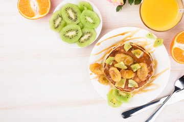 Homemade breakfast on a wooden white table. Stack of pancakes with a grilled bananas and a caramel. Glass of juice, orange, kiwi slices. Healthy breakfast concept, flat lay, top view