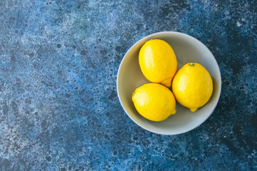 Three ripe yellow lemons in a grey bowl on a blue stone nackground. Top view and copy space.
