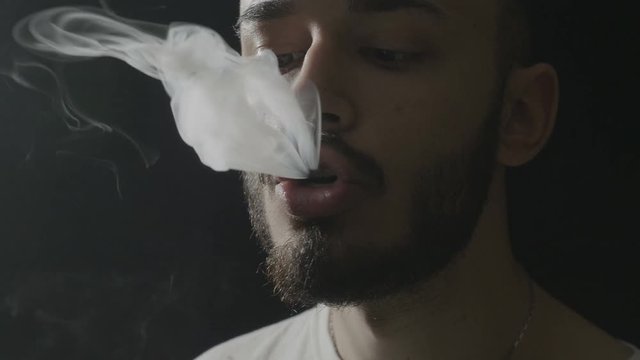 Close up of a young hipster man with beard inhaling from a vaporizer and exhaling large amount of smoke while doing vaping tricks