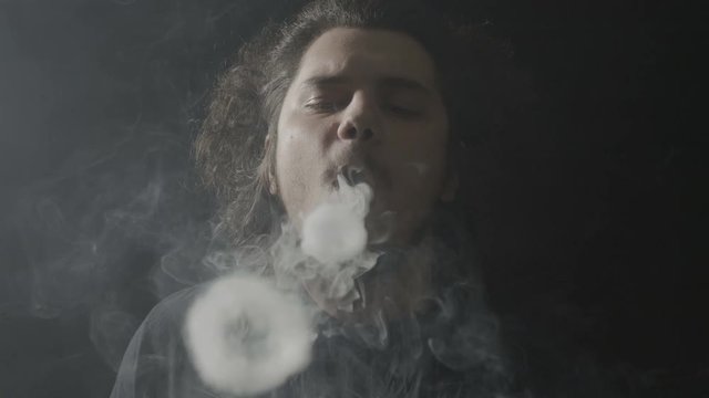 Portrait of a an adult vaper with long hair performing impressive vaping steam circle tricks after inhaling from an electronic cigarette