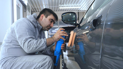 A man in a special suit polishes a gray car body, a tool for polishing cars, into a workshop.