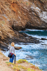A girl photographer with a tripod in her hand, stands on the shore of the bay against the background of the sea and red rocks.