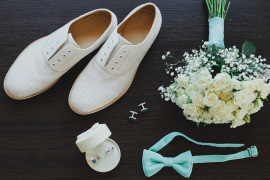 Leather white groom wedding shoes on the wooden background in sun rays. Rings, bow tie and cufflinks with elegant bouquet. Elegant wedding details flat lay. Tiffany light blue color.