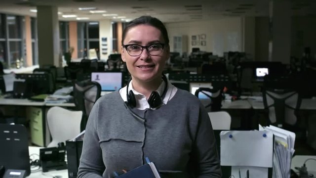 PAN of mature female call centre manager in glasses and with headset around her neck holding planner and looking at camera in empty office