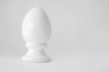 Single white decorative ceramic statuette egg in eggcup on gray white blurred background with copy-slace left. Easter religious Christian symbol.