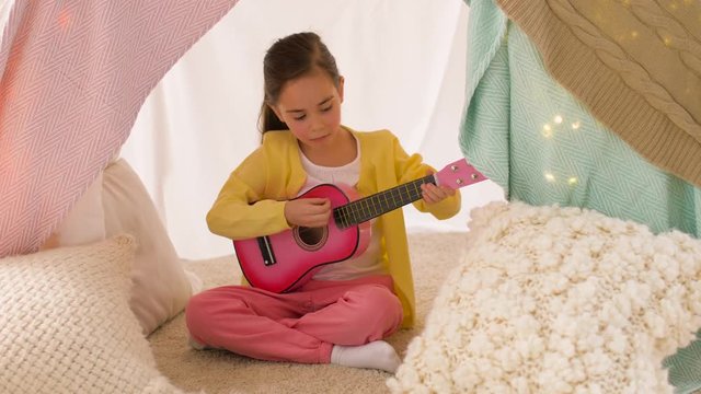 childhood and hygge concept - happy little girl with toy guitar playing music in kids tent at home