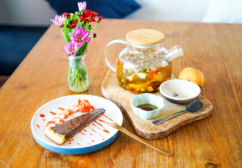 A piece of chocolate cake with strawberries lies on a white plate, sprinkled with jam. A large teapot of green Chinese tea is on the table with a wooden backdrop. Side view with copy space 