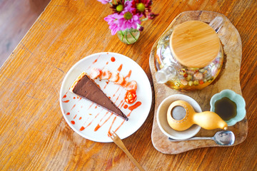 A piece of chocolate cake with strawberries lies on a white plate, sprinkled with jam. A large teapot of green Chinese tea is on the table with a wooden backdrop. Top view with copy space 