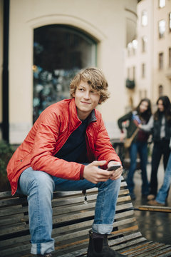Full length of teenage boy holding smart phone while sitting on bench with friends in background