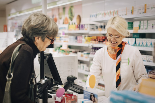 Smiling female owner standing with senior customer at checkout in pharmacy store