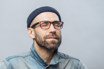 Portrait of relaxed unshaven male with closed eyes wears glasses, keeps eyes closed, enjoys calm atmosphere, isolated over white background. People, relaxation and facial expressions concept - Powered by Adobe