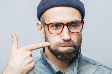 Close up shot of unshaven male with thick beard and mustache blows cheeks and indicates with fore...