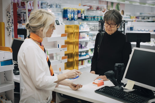 Female owner holding prescription paper with senior customer standing at checkout in pharmacy store