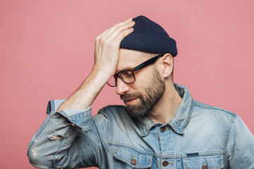 Horizontal portrait of stressful stylish unshaven male regrets something, keeps hand on head, looks...