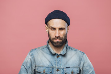 Photo of good looking bearded male blinks eye and looks seriously into camera, wears denim jacket...