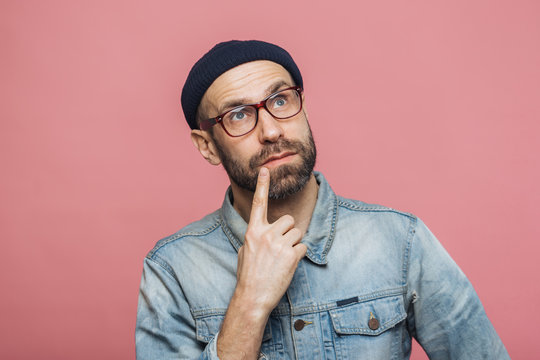 Thoughtful bearded male being deep in thoughts, looks with pensive expression into camera, concentrated on something, poses against pink background. People, facial expressions, daydreaming concept