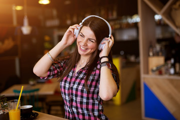 Young cheerful beautiful girl is enjoying the music on her headphones while sitting at the table in the coffee shop and drinking juice.