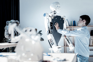 It looks so real. Side view on an impressed schoolboy grinning broadly while standing in front of a human like robot and touching it for the first time.