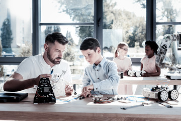 Help is what really matters. Serious mature guy in casual sitting next to his smart student and discussing the nuts and bolts of their technical creation and adjusting wires in a workshop.