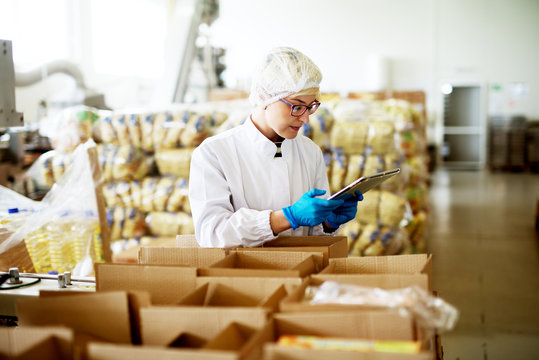 Young beautiful worried female worker in sterile clothes is using a tablet while being leaned against the stacks of open boxes inside of factory storage.