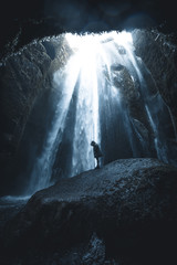 Women standing in jacket on a rock in front of waterfalls in a cave with incoming sunlight in Iceland
