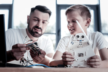 Which one to choose. Selective focus on colorful wires and a white robot held by smart mature man and his adorable student sitting in a workshop and constructing new machine together.