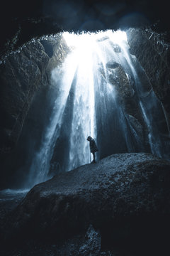 Women standing in jacket on a rock in front of waterfalls in a cave with incoming sunlight in Iceland