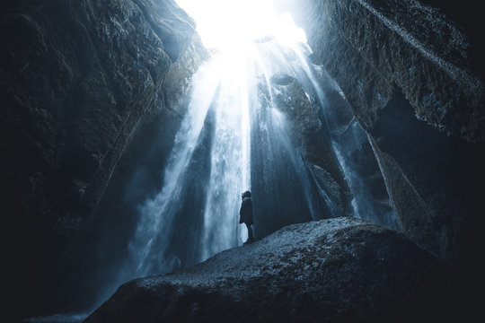 Women standing in jacket on a rock in front of waterfalls in a cave with incoming sunlight in Iceland