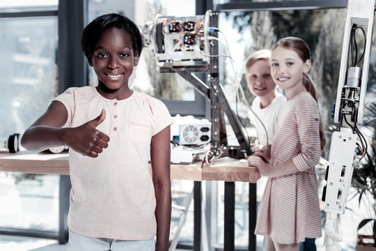 What a cool experience. Selective focus on a cheerful schoolgirl looking into the camera with a smile on her face and thumbing up after examining a robotic machine with her friends.