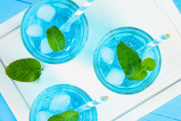 Blue cocktail with ice and mint in glasses on a white wooden board on a blue table. Top view.