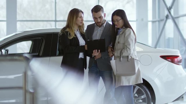 Tilt up of professional auto saleswoman in formal clothing holding clipboard and talking with young couple choosing new car at dealership