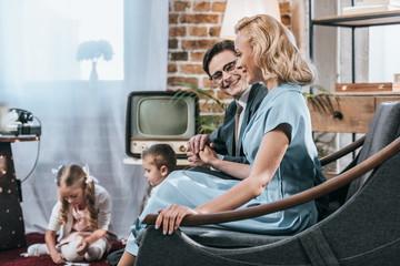 happy old-fashioned parents sitting on sofa and holding hands while children playing with domino...