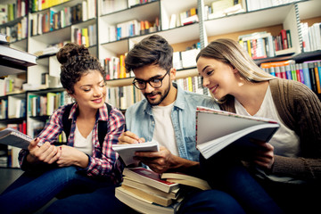 A group of young focused joyful students studying, explaining each other and writing notes in a notebook while sitting on a floor in front of a library bookshelf.