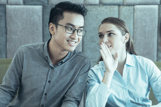 Content middle-aged business woman whispering secret to male colleague sitting next to her at table at meeting. There is another woman seen partly.