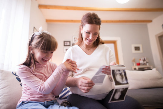 Beautiful pregnant mother and little adorable daughter looking excited at the ultrasound pictures of the baby while sitting on the sofa at home.