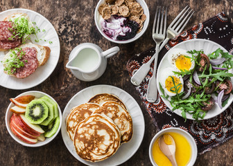 Greek yogurt with whole grain cereals and berry sauce, pancakes, arugula, cherry tomatoes, boiled eggs salad, kiwi, apples fruit, salami and cream cheese sandwiches on a wooden background, top view
