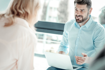 Fototapeta premium Emotional discussion. Handsome male keeping smile on face and holding laptop on knees, being in all ears