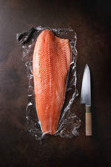 Whole raw uncooked salmon fillet on food film with chef's knife over dark brown texture background. Top view, space.